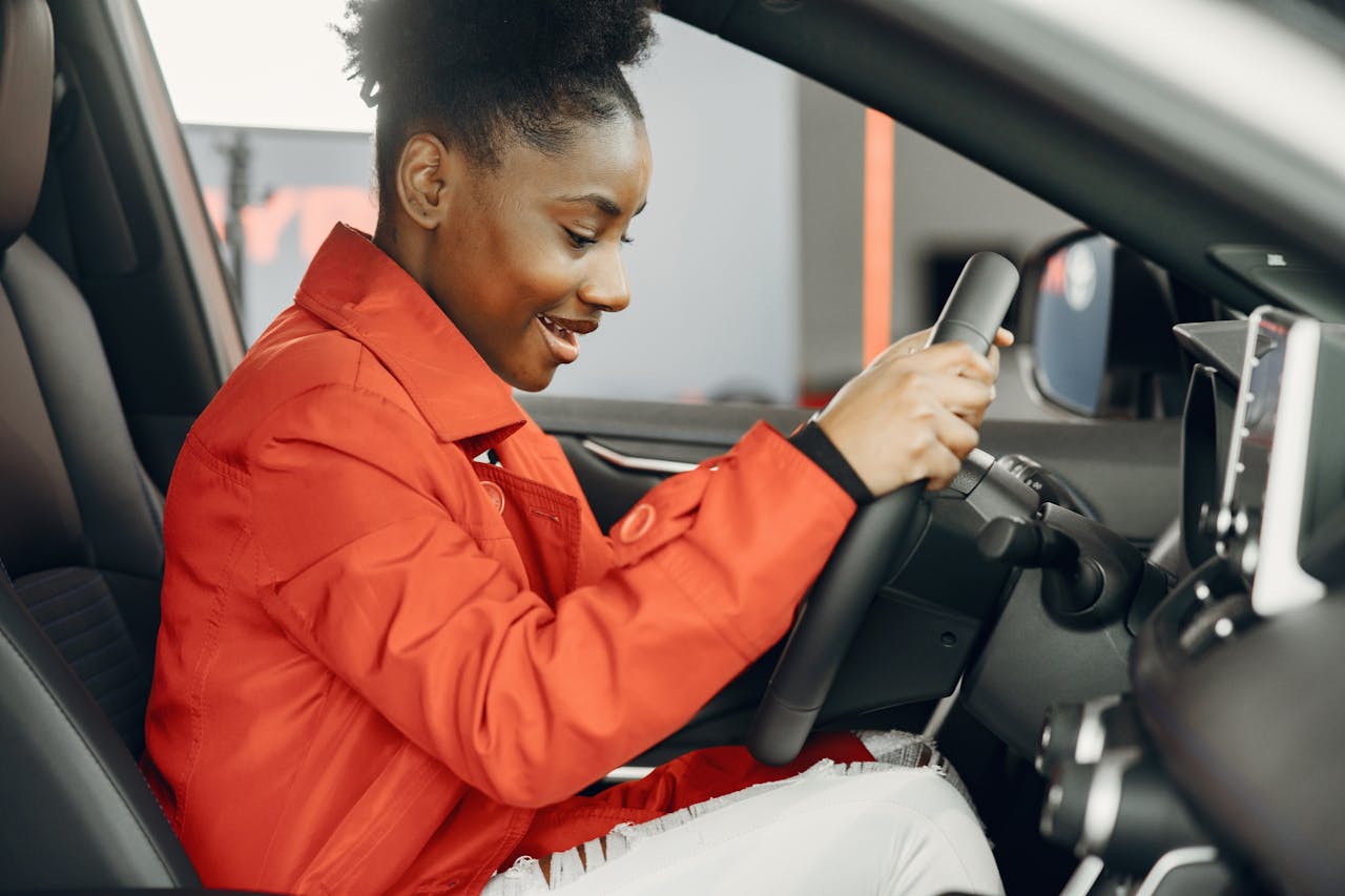 services-01 Young woman in an orange jacket enthusiastically trying out a car's steering wheel.